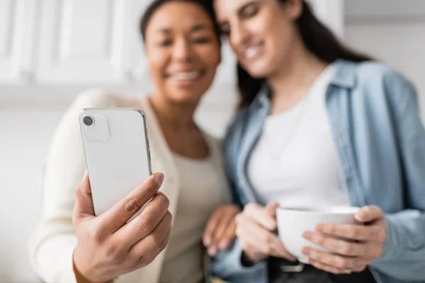 Mujer multirracial sosteniendo teléfono inteligente cerca de pareja lesbiana con taza de café - foto de stock