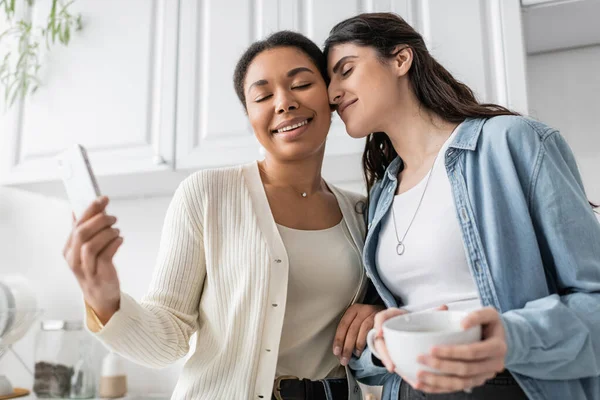Joyful multiracial woman holding smartphone near lesbian partner with cup of coffee — Stock Photo