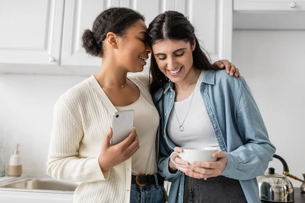 Joyful multiracial woman holding smartphone and hugging lesbian partner with cup of coffee — Stock Photo