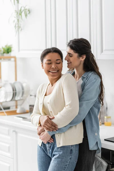 Весела лесбіянка обіймає багаторасову дівчину на сучасній кухні — стокове фото