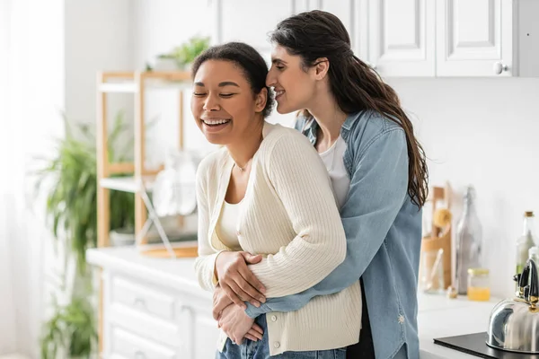 Mulher lésbica positiva abraçando namorada multirracial na cozinha moderna — Fotografia de Stock