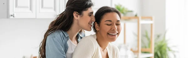 Fröhliche lesbische Frau umarmt multirassische Freundin in der modernen Küche, Banner — Stockfoto