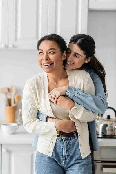 Весела лесбіянка обіймає позитивну багаторасову дівчину на сучасній кухні — стокове фото