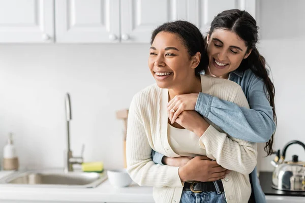 Fröhliche lesbische Frau umarmt freudige multirassische Freundin in der modernen Küche — Stockfoto