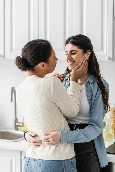 Pleased lesbian woman hugging with multiracial girlfriend in modern kitchen — Stock Photo
