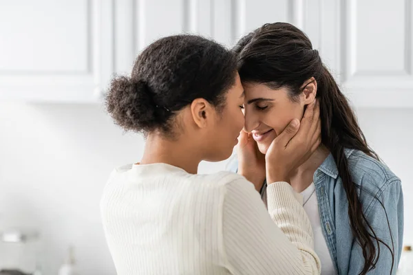 Curly multiracial woman hugging happy girlfriend with closed eyes at home — Stock Photo