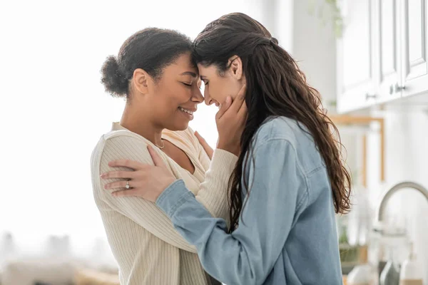 Curly multiracial woman hugging cheerful girlfriend with closed eyes at home — Stock Photo