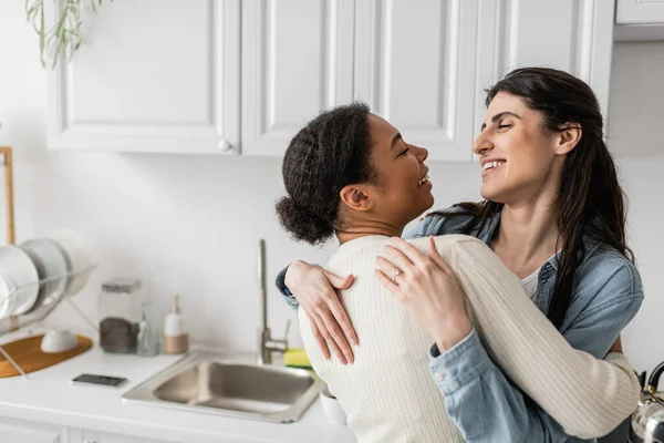 Femme lesbienne ravie souriante et étreignant petite amie multiraciale positive avec les cheveux bouclés — Photo de stock