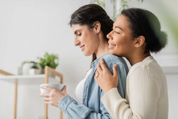 Overjoyed multiracial woman with engagement ring on finger hugging girlfriend holding cup of coffee — Stock Photo