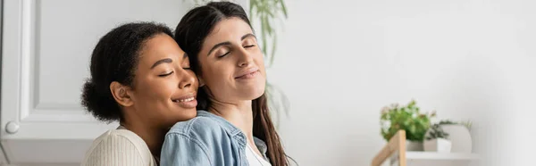 Cheerful multiracial lesbian woman with curly hair hugging girlfriend at home, banner — Stock Photo