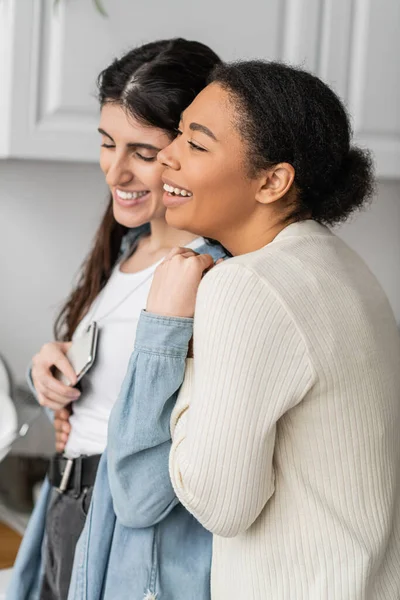 Überglückliche lesbische Frau hält Smartphone und umarmt glückliche multirassische Freundin — Stockfoto
