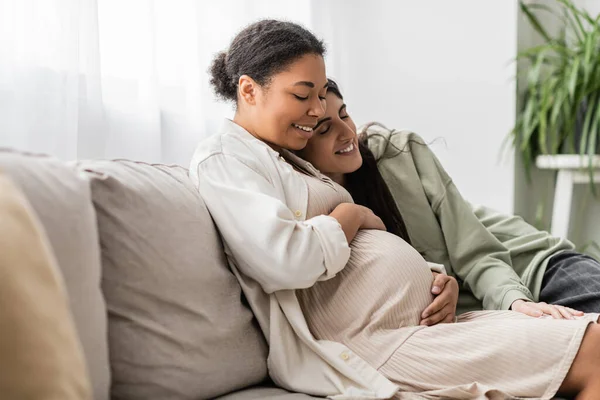 Glücklich lesbische Frau umarmt schwangere multirassische Frau, während sie auf dem Sofa sitzt — Stockfoto