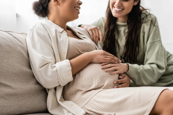 Обрізаний вид щасливої лесбіянки, обіймаючи вагітної багаторасової дружини, сидячи на дивані — стокове фото