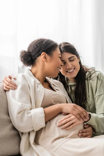 Fröhliche lesbische Frau umarmt schwangere multirassische Frau, während sie auf dem Sofa sitzt — Stockfoto
