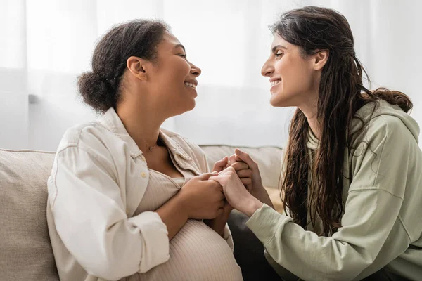 Позитивная лесбиянка, держащаяся за руки с беременной многорасовой женой, сидя на диване — стоковое фото