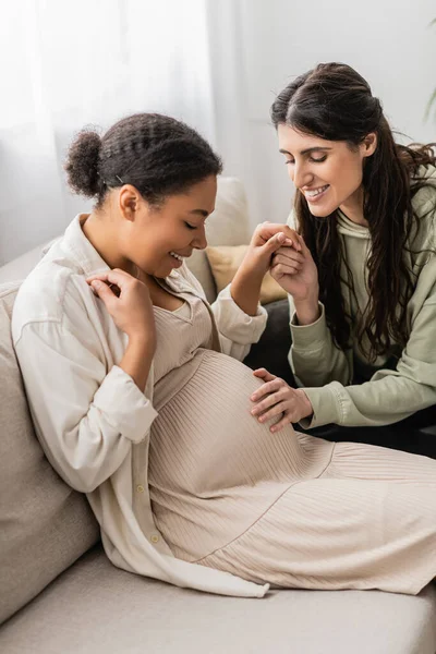 Весела лесбіянка торкається живота вагітної багаторасової дружини, сидячи на дивані — стокове фото
