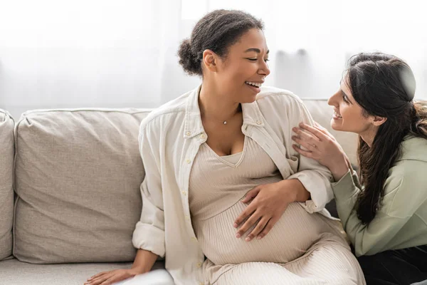 Feliz lesbiana mujer mirando alegre embarazada multirracial esposa mientras sentado en sofá - foto de stock