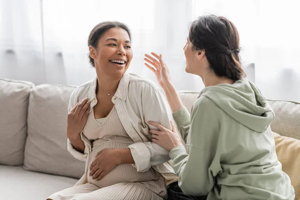 Happy lesbian woman chatting with cheerful pregnant multiracial wife while sitting on sofa — Stock Photo