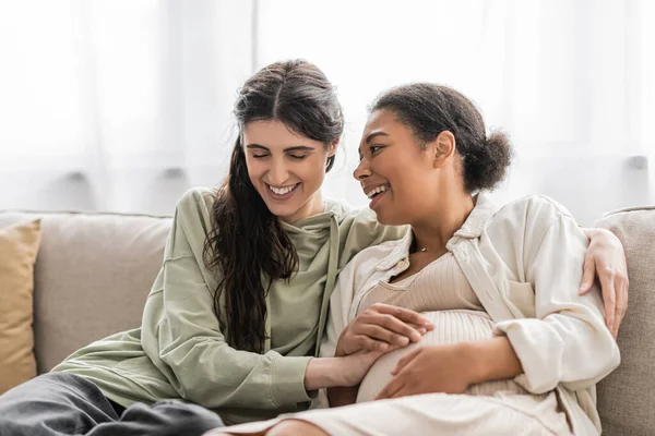 Mujer lesbiana llena de alegría abrazando embarazada esposa multirracial mientras se sienta en el sofá - foto de stock