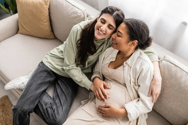 Високий кут зору щасливої лесбіянки, обіймаючись вагітною багаторасовою дружиною, сидячи на дивані — стокове фото