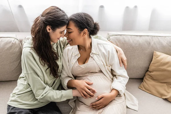 Щаслива лесбіянка обіймає вагітної багаторасової дружини, сидячи на дивані — стокове фото