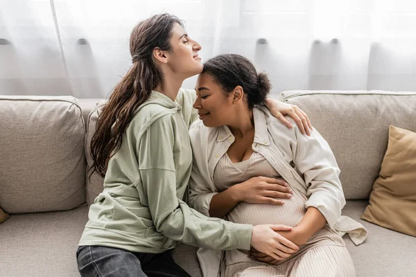 Alegre lesbiana abrazando alegre embarazada esposa mientras sentado en sofá - foto de stock