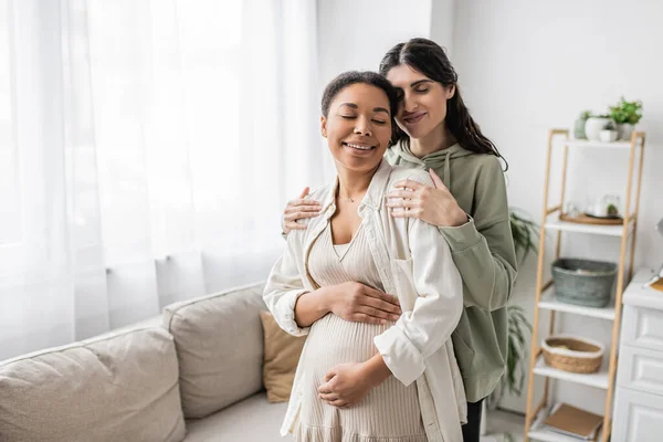 Feliz mulher lésbica sorrindo enquanto abraça a esposa multirracial grávida na sala de estar — Fotografia de Stock
