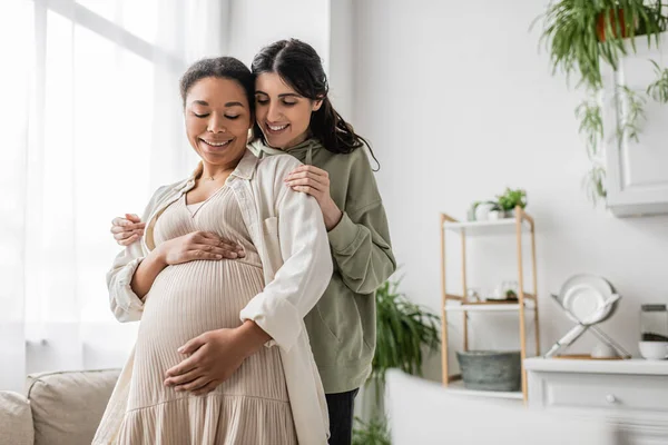 Mulher lésbica alegre sorrindo enquanto abraça a esposa multirracial grávida na sala de estar — Fotografia de Stock