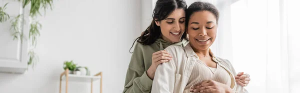 Feliz mujer lesbiana sonriendo mientras abraza esposa alegre en sala de estar, pancarta - foto de stock