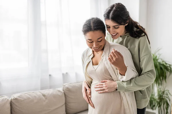 Fröhliche lesbische Frau lächelt, während sie den Bauch ihrer schwangeren multirassischen Frau im Wohnzimmer umarmt — Stockfoto