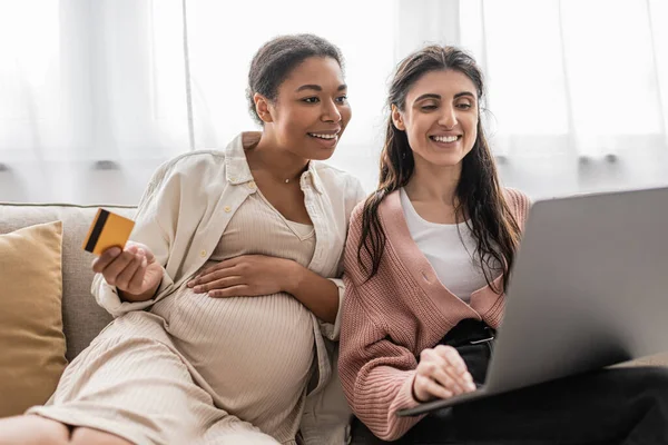 Embarazada multirracial mujer celebración de la tarjeta de crédito, mientras que hacer compras en línea con feliz pareja lesbiana - foto de stock