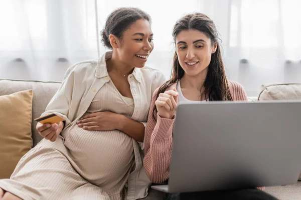 Embarazada mujer multirracial celebración de la tarjeta de crédito mientras que hace compras en línea con alegre pareja lesbiana - foto de stock