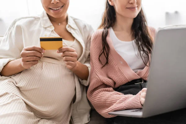 Обрезанный вид беременной многорасовой женщины, держащей кредитную карту во время покупок в интернете с веселым лесбийским партнером — стоковое фото