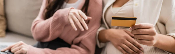 Частковий вид вагітної багаторасової жінки, що тримає кредитну картку біля лесбійського партнера, банер — стокове фото