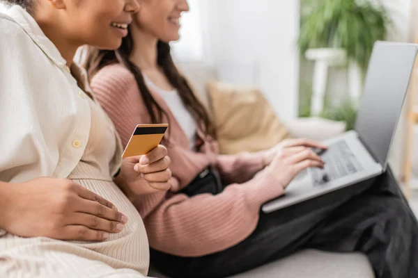 Vue partielle de la femme enceinte heureuse et multiraciale tenant la carte de crédit tout en faisant des achats en ligne avec un partenaire lesbien — Photo de stock