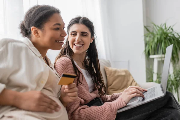 Feliz lesbiana mujer mirando embarazada multiracial pareja mientras haciendo compras en línea juntos - foto de stock