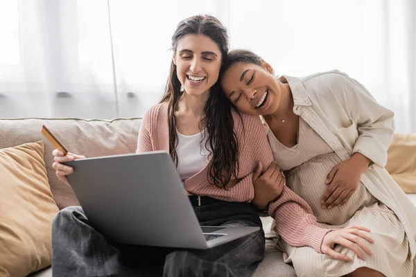 Щаслива лесбіянка тримає кредитну картку під час онлайн-покупки біля вагітного багаторасового партнера — стокове фото