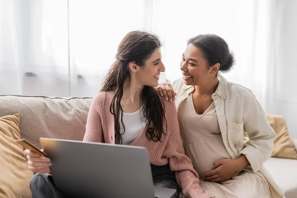 Happy lesbian woman holding credit card while doing online shopping with cheerful and pregnant multiracial partner — Stock Photo