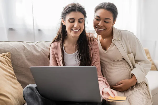 Alegre y embarazada mujer multirracial mirando a la computadora portátil mientras hace compras en línea con feliz pareja lesbiana - foto de stock