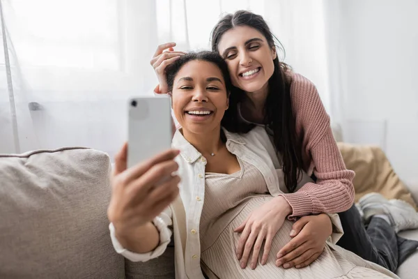 Femme multiraciale heureuse et enceinte tenant smartphone tout en prenant selfie avec partenaire lesbienne — Photo de stock