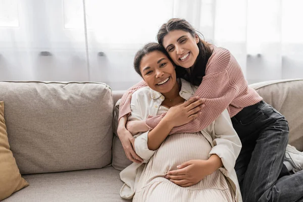 Feliz lesbiana mujer abrazando embarazada multirracial pareja y sentado en sofá - foto de stock