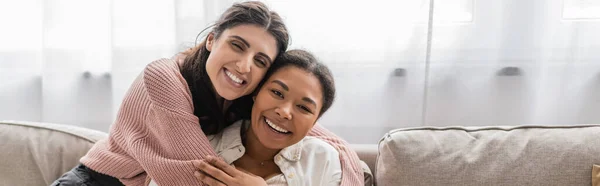 Glückliche lesbische Frau umarmt multirassischen Partner und sitzt auf Couch, Banner — Stockfoto