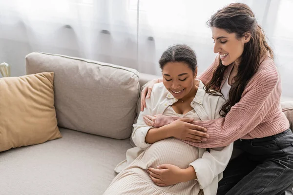 Gioiosa donna lesbica abbracciare incinta multirazziale partner e seduto sul divano — Foto stock