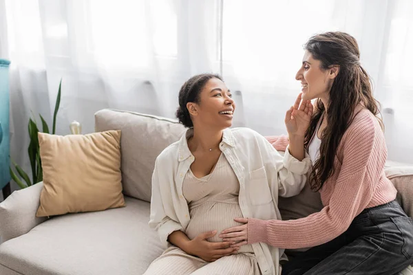 Положительная лесбиянка обнимает беременную многорасовую партнершу и сидит на диване — стоковое фото