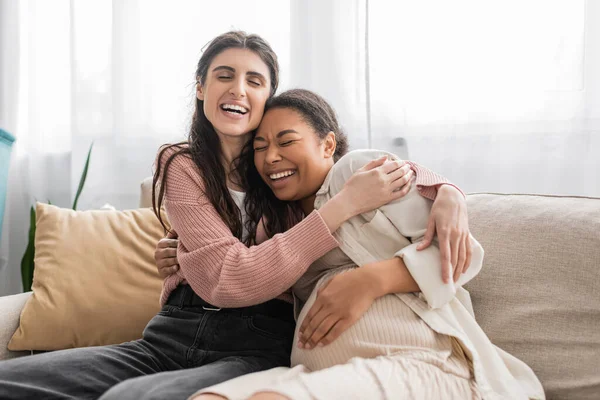 Aufgeregte lesbische Frau umarmt schwangere multirassische Partnerin und sitzt auf Couch — Stockfoto