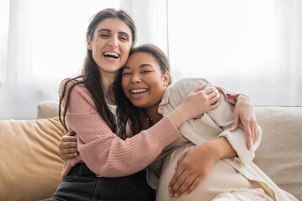 Overjoyed lesbian woman hugging pregnant multiracial partner and sitting on couch — Stock Photo
