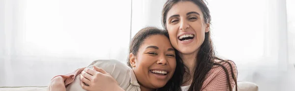 Задоволена лесбіянка обіймає багаторасову дівчину у вітальні, банер — стокове фото