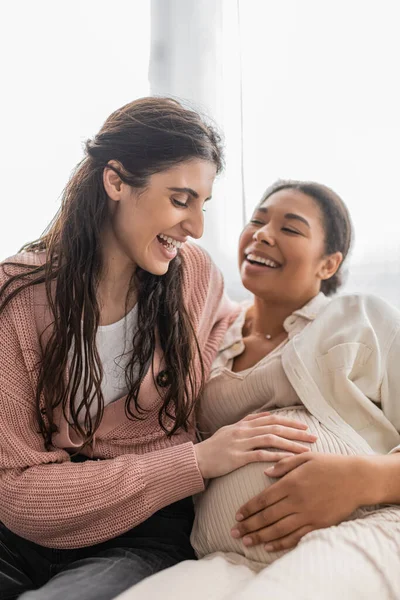 Overjoyed lesbian woman touching belly of pregnant multiracial partner and sitting on couch — Stock Photo