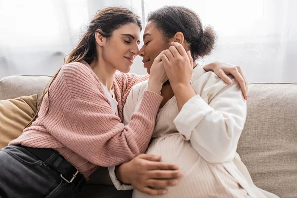 Happy lesbian woman holding hands with pregnant multiracial partner and sitting on couch — Stock Photo