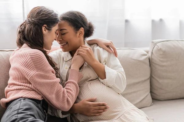 Позитивна лесбіянка обіймає вагітного багаторасового партнера і сидить на дивані — стокове фото